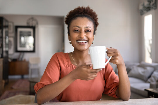 Portrait of cheerful African woman sitting at table, holding white mug and smiling with warmth and joy at camera, enjoying weekend at home and moments of comfort and relaxation with favourite beverage