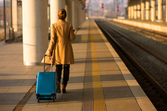A woman in a tan coat pulls a blue rolling suitcase while checking her phone at a modern train station, heading toward her departure platform.