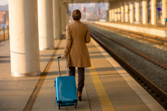 Back view of a woman in a tan coat pulling a blue rolling suitcase along a sun-drenched railway platform, symbolizing departure and the start of a new journey.