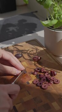 Hand slices black olives on a wooden cutting board near a potted herb. Concept of cooking, appetizer and fresh ingredient prep