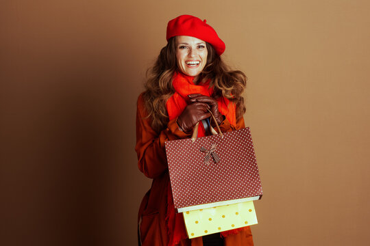 Joyful woman with wavy brown hair, red beret, orange scarf, and brown coat, smiles holding two patterned shopping bags. Warm background embodies autumn satisfaction and stylish retail therapy.