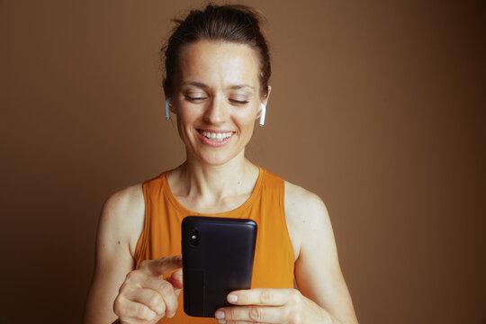 A smiling woman in an orange tank top and earbuds looks down, engaging with her smartphone, depicting casual tech interaction and personal entertainment.