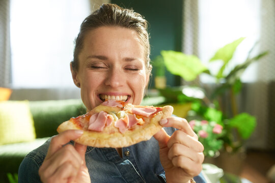A close-up of a young woman with her eyes closed in contentment, holding a slice of ham and cheese pizza with both hands in a bright living room.