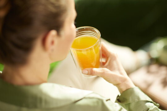 Close-up view from behind a woman in an olive-green shirt as she prepares to drink a glass of orange juice, highlighting a moment of self-care and morning nutrition.