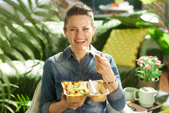 A woman with a bun smiles while holding a partitioned tray of fresh fruit, surrounded by greenery in a sunlit home interior, promoting healthy food choices.