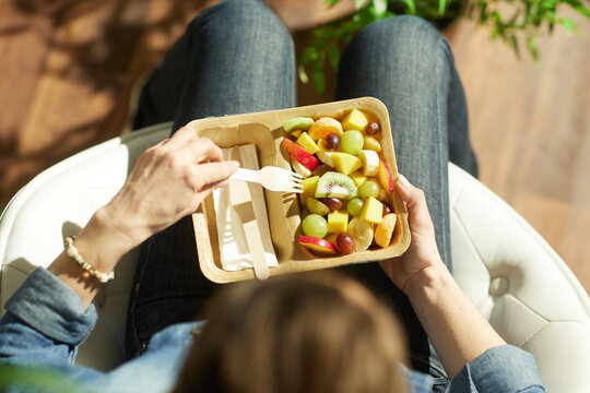 An adult woman in a denim shirt sits in a white chair, enjoying a fresh fruit salad with kiwi, grapes, and bananas from a partitioned cardboard container in a bright room.