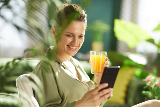 A woman with her hair in a bun smiles while looking at her phone and holding a glass of orange juice, surrounded by vibrant houseplants.