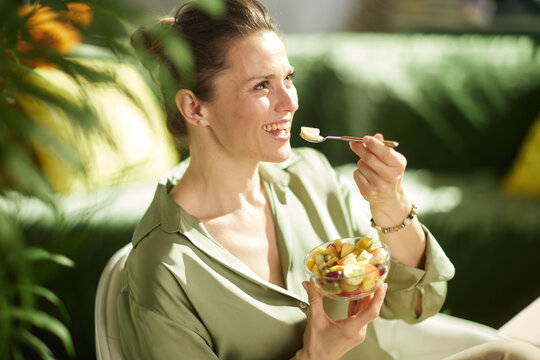 A cheerful woman enjoys a fresh bowl of banana and kiwi salad, captured in warm natural light with a lush, blurred green background, emphasizing wellness and joy.