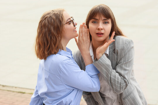 Gossip and secret sharing, two young women whispering confidential information with surprised reaction outdoors