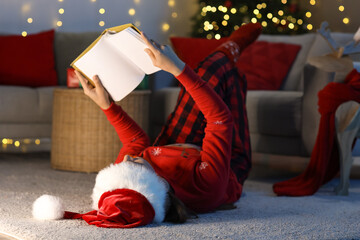Fototapeta premium Young girl in Santa hat reading book at home on Christmas eve