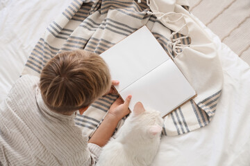 Fototapeta premium Little boy with cute cat reading book on bed, top view