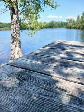 Old wooden pier beside a tranquil lake with green trees. Landscape of a boardwalk with worn planks and shadows beside a blue lake with trees on the distant shore. Canadian landscape.
