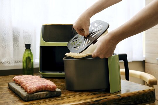 Person places parchment paper under crisper tray in food basket of air fryer in kitchen. Fresh sausage sits on wooden cutting board. Olive oil bottle present on table next to air fryer