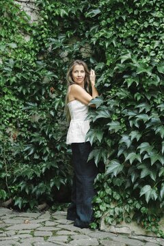 Middle-aged woman with long hair poses next to wall covered in green ivy in urban area. Sunlight shines bright, highlighting green plants around. She has natural stance and smiles gently