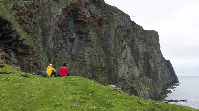 A couple sitting below the towering cliffs of the Isle of Foula, Shetland, Scotland, United Kingdom