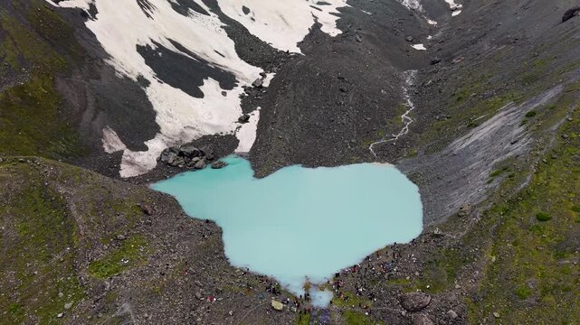 Overhead drone shot hovering above a bright turquoise mountain tarn surrounded by rocky scree slopes, patches of melting snow, and a group of tiny hikers gathered along the shore.