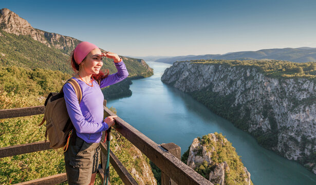 Smiling female hiker shades her eyes at a scenic overlook above a sweeping turquoise Danube river Iron Gates gorge, full of wonder and wanderlust. Adventure concept.