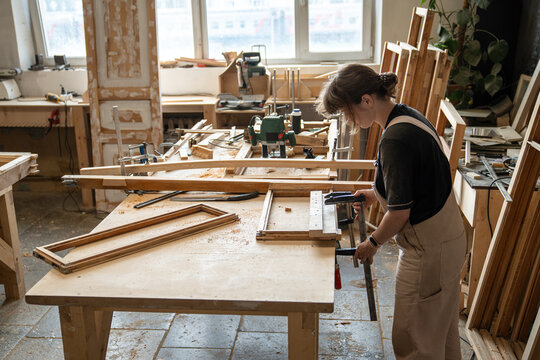Woman crafter in restoration workshop fixating parts of vintage wooden window frame using clamps, female carpentry employee working in woodshop at workbench with timber materials and hardware tools.