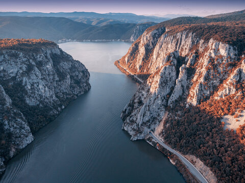 Aerial view of the Danube river passing between steep rocky cliffs of Iron Gate and forested mountains during a colorful sunset at autumn