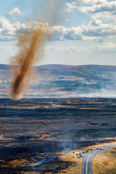 Tall vortex of smoke and dust rising from smouldering black ground after hill fire, spectators and vehicles gathered along rural road in summer