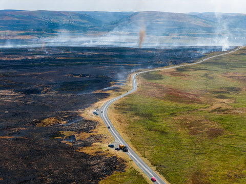 Smoke whirl forming above burnt heathland after summer wildfire, vehicles parked along road dividing charred landscape from green moor, South Wales