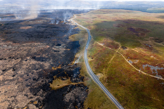 Smoke rising from charred ground after a huge summer wildfire, black scorched earth halted by a tarmac road dividing burned peat from undamaged moorland hillside