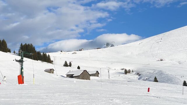 Wide view of a snowy ski slope in Megeve, Haute-Savoie, French Alps, featuring traditional wooden chalets, ski lift infrastructure, scattered evergreen trees, and a partly cloudy blue sky.