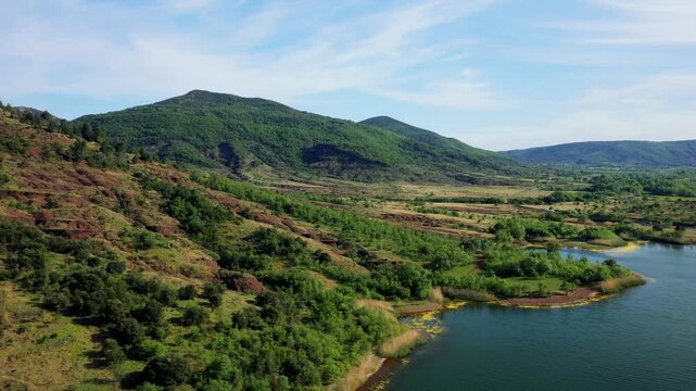 Aerial footage of the shoreline of Lac de Salagou in Herault, Occitanie, France. The video shows the lake's banks with distinctive red earth, lush green vegetation, and rolling hills under a blue sky.