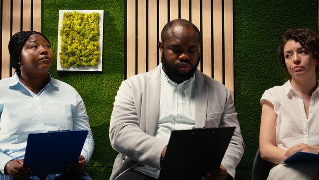 Diverse candidates await job interviews in an office hallway, showing anticipation while preparing information for the recruitment process.