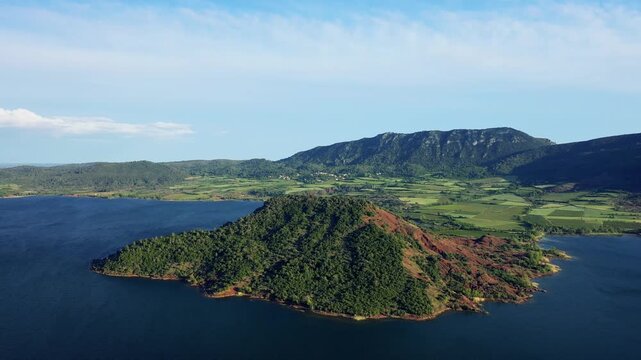 Aerial footage of a green peninsula jutting into the blue waters of Lac de Salagou, surrounded by rolling hills, fields, and distant mountains under a clear sky.