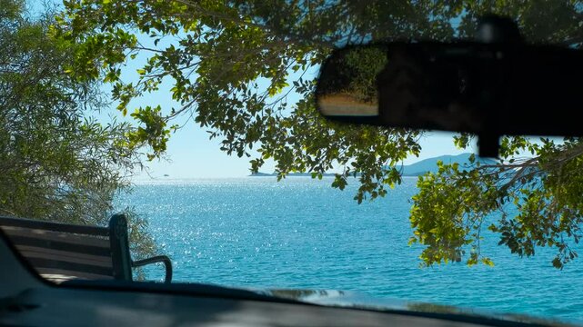 Vehicle parked near coast enjoying stunning ocean view. Sunlight reflects on a calm, blue ocean, seen through the windshield of a parked vehicle near a leafy tree and a bench, offering a coastal view