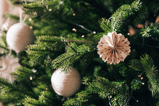 Christmas tree with paper ornament hanging on spruce branch with shining garland with glowing lights and decorations background, capturing magical spirit of winter holidays and festive home decor.