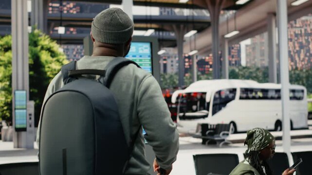 Passenger carrying baggage arrives in a coach terminal for an intercity route and waits for a bus connection, pulling a trolley and backpack toward the seating area benches before departure.