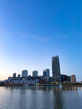 City skyline by the water at dusk. Buildings stand by the water as the sky turns blue at dusk. The lights reflect on the calm water surface.
