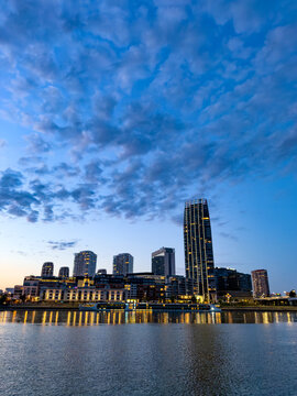 Modern skyline over calm river at twilight. Scattered clouds drift above tall buildings while night lights reflect on water.
