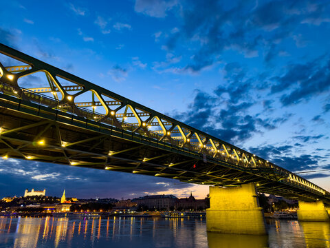 Illuminated steel truss bridge over river at night with castle. Yellow lights glow along structural beams while castle stands on distant hill under dusk sky.
