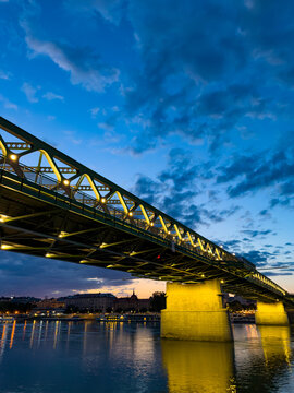 Bridge lit up at dusk by the river. A bridge with lights shines over the river during dusk. Clouds fill the sky as the day ends in the city.
