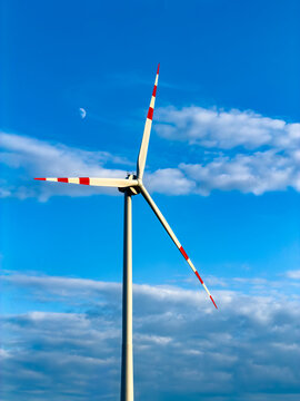 Wind turbine against blue sky and clouds. A wind turbine stands tall in the blue sky with clouds around and a small moon visible in the background during the day.