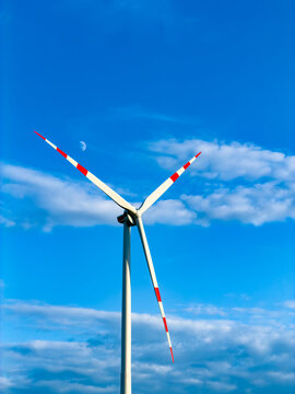 Single wind turbine against clear blue sky with crescent moon. Red tipped propeller blades stand out against soft clouds in vertical composition.