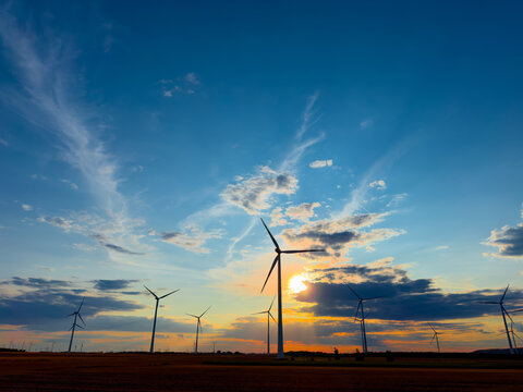 Wind turbines on agricultural field at golden sunset. Sunlight breaks through dark clouds over silhouetted power generators during dusk.