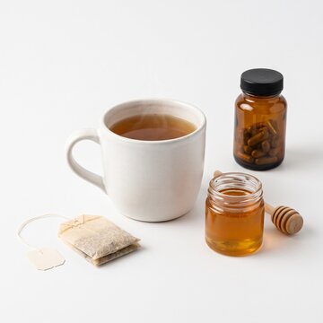 A cup of hot herbal tea with a tea bag, a small jar of honey, and a bottle of pills on a white surface. The tea is steeping and emitting steam, suggesting a soothing and healing beverage.