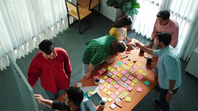 Top view of diverse business people writing and stick sticky notes at whiteboard. Creative team working together and brainstorming marketing idea for startup project at table with notes. Convocation.