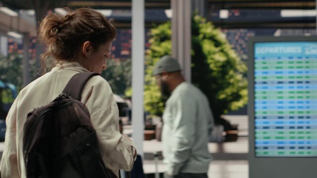 Woman walks through a bus station with a trolley and backpack, carrying baggage into the coach terminal waiting area before sitting on a metal bench.