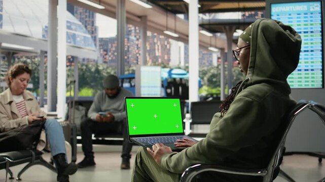 Tourist uses a green screen laptop in a bus station during long-distance travel, typing on the chroma key notebook keyboard to complete work assignments while waiting at the coach station.