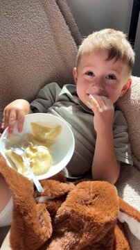 Happy little boy lying down back and eating delicious homemade dumplings with sour cream, relaxed cute child enjoying food on cozy sofa