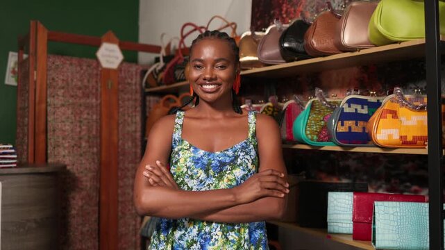 African woman small business owner smiling with arms crossed in her retail store selling handbags, SME
