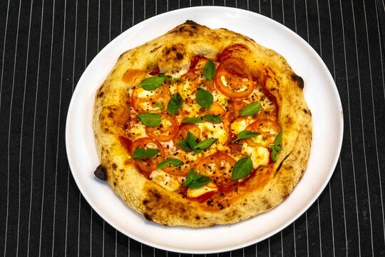 Neapolitan-style personal pizza with naturally leavened long-fermentation dough, sliced tomatoes, melted mozzarella, fresh basil and oregano, served on a white plate over a dark pinstriped cloth.