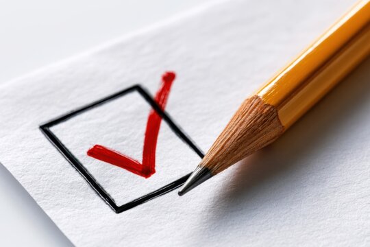 Yellow pencil resting on paper with a bold red checkmark inside a black square box indicating completion or approval on a form or checklist