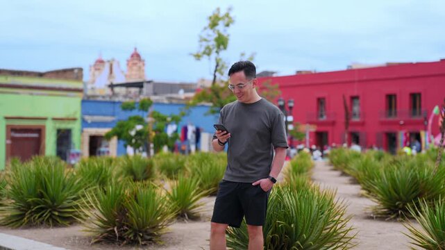Young latin man happily using a smartphone while walking through a beautiful park with agave plants in oaxaca, mexico, with colorful colonial buildings and a church in the background