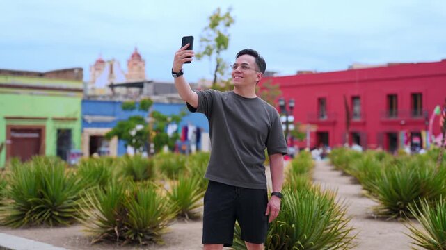 Joyful latin tourist taking a selfie with his smartphone while visiting a colorful plaza in oaxaca, mexico, sharing his travel adventures on social media with a smile on his face